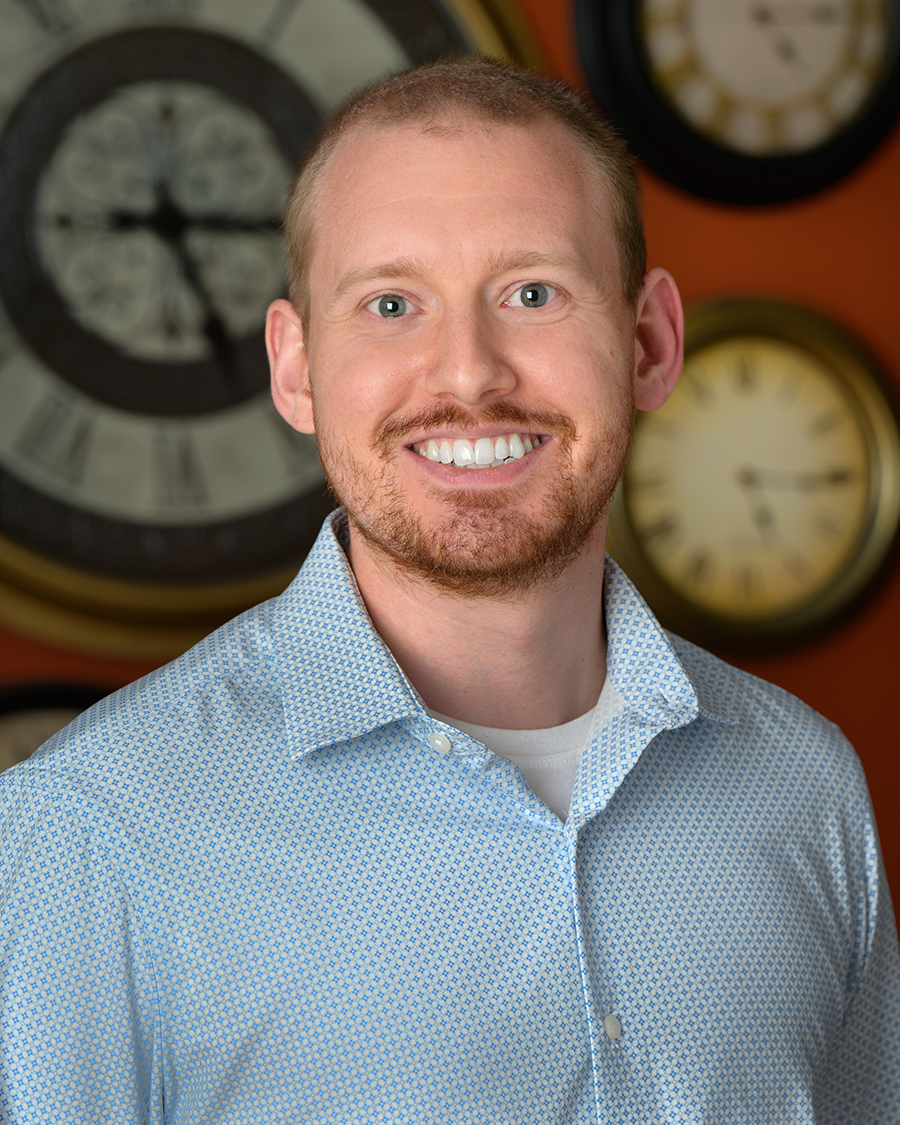 A smiling man with a light beard wearing a blue patterned button-down shirt, with several analog clocks hanging on the wall behind him.
