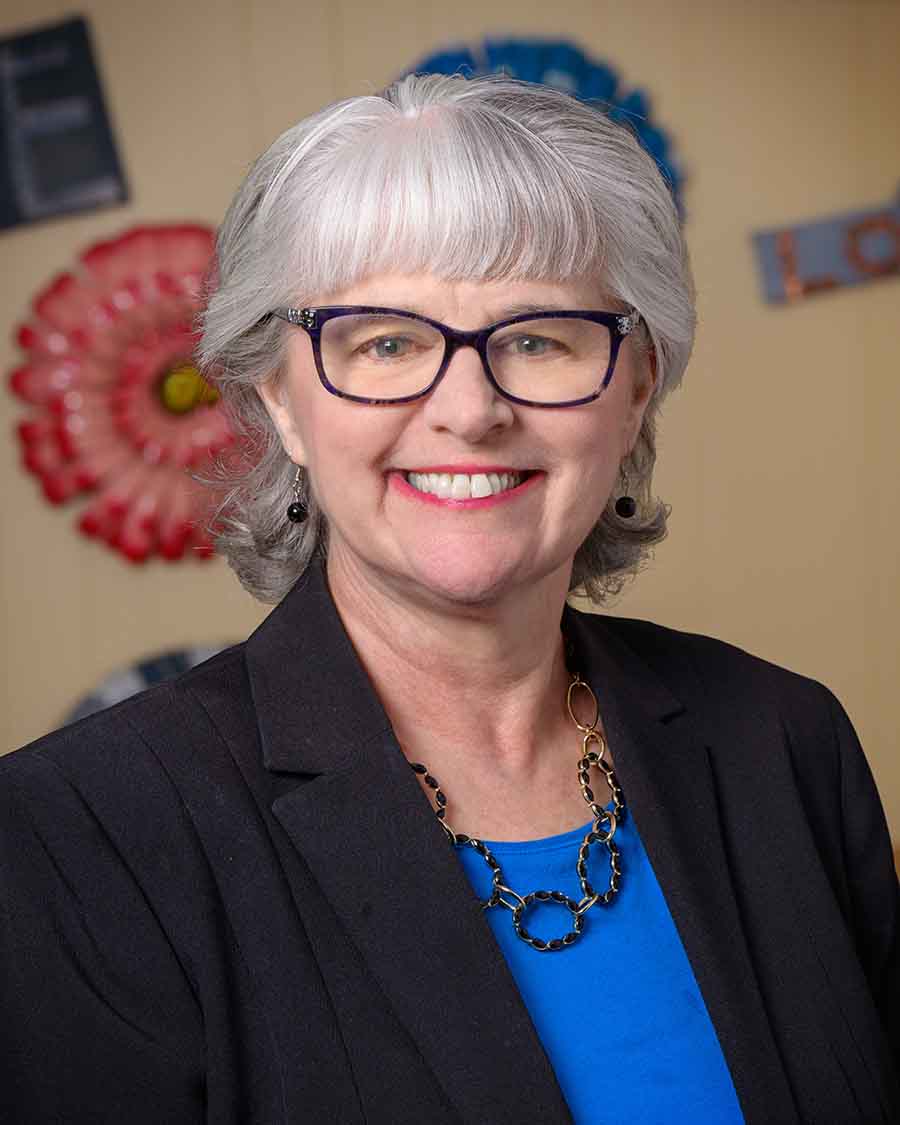 A smiling woman with gray hair and glasses wearing a blue top, black blazer, and a linked necklace.